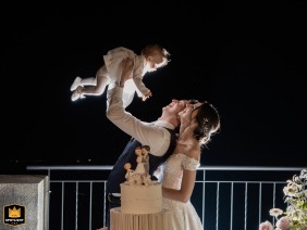 A joyful family portrait captured at Restaurant Vetta in Monte Bre, Lugano, Switzerland, showcasing the bride and groom lifting their baby overhead during the night cake cutting on their wedding day.